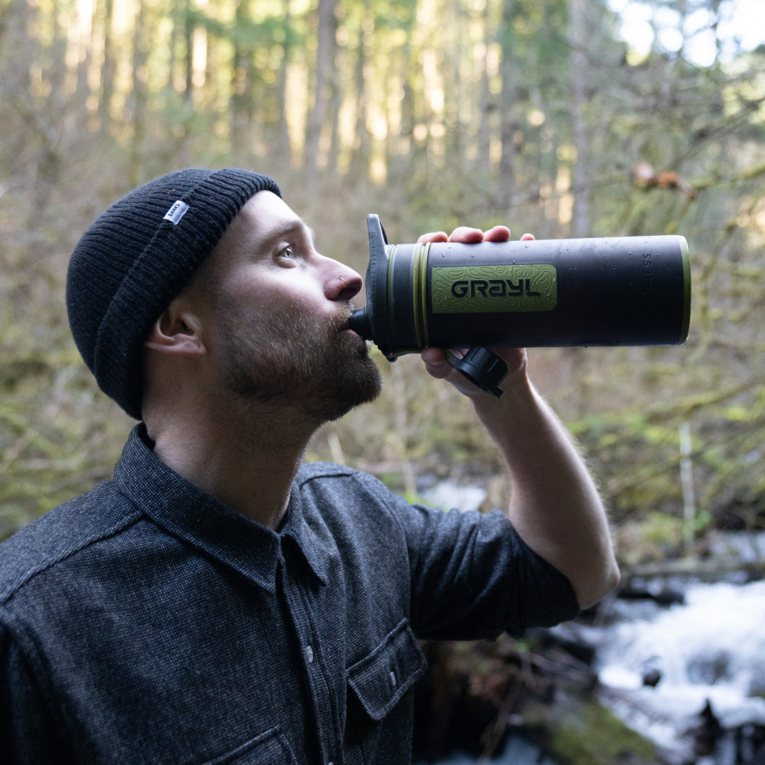 hombre bebiendo de su Grayl con un arroyo forestal al fondo